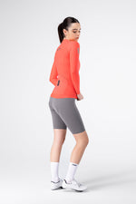 Female model wearing Gobik Avalon long sleeve jersey in lava with gray cycling shorts, side view