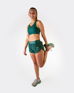 Woman wearing forest green ChicknLegs sports bra and matching shorts while stretching
