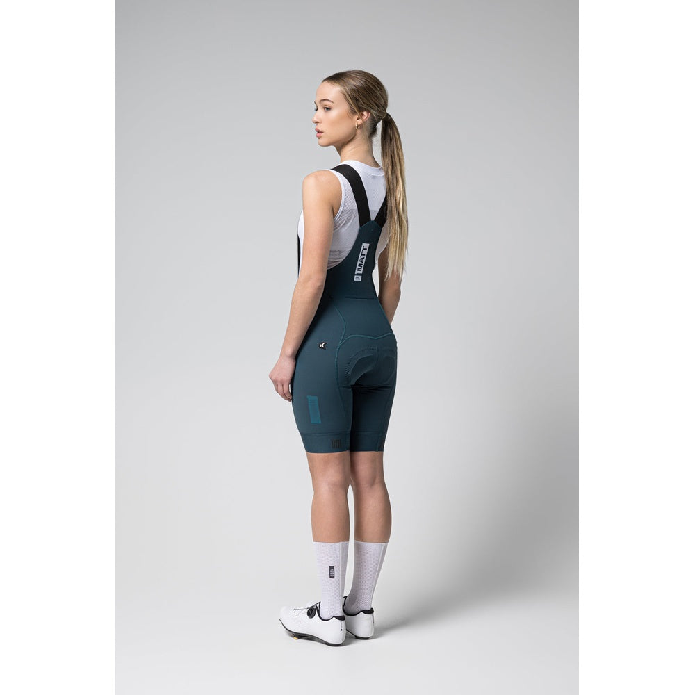 Woman wearing dark teal Gobik Matt 2.0 bib shorts with suspenders, back view