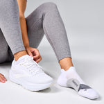 Person tying white Brooks running shoes wearing white Feetures ankle socks on gray background