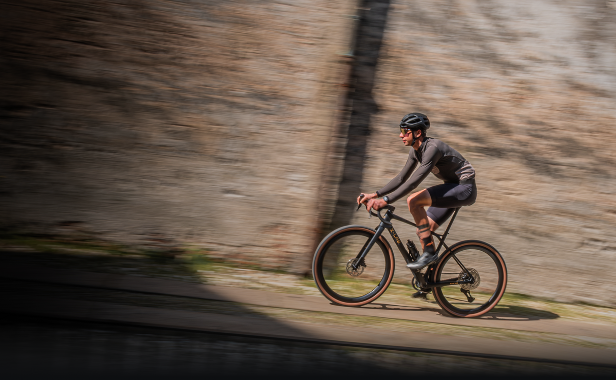Cyclist riding X-LAB GT8 gravel bike in black colorway along stone wall at speed